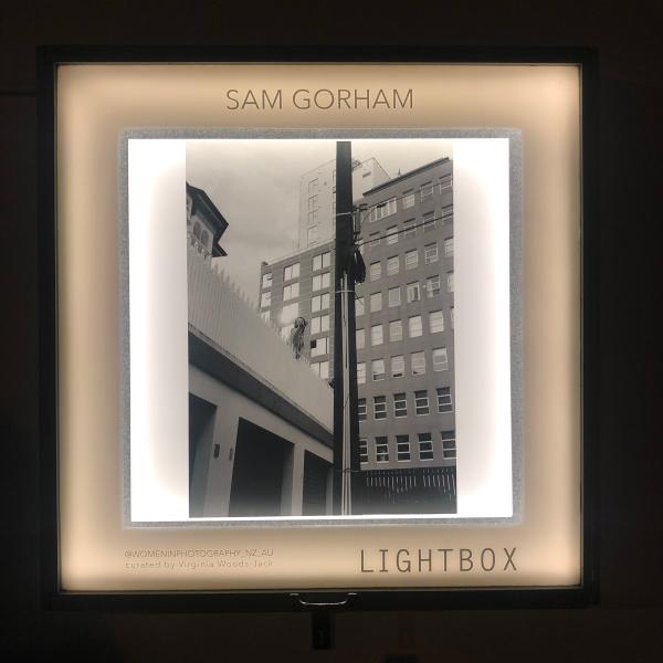 Image of lightbox at Thistle hall lit up at dusk with Black and white image of young woman reaching out to the photographer in a gesture of friendship. Sun burst on window of building behind subject. 