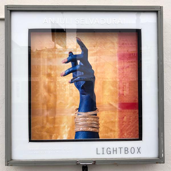 Install shot of artwork in the lightbox at thistle hall - Woman's hand and lower arm painted bright blue with long pink glittered nails. Multiple gold bangles. Gesture of hand is loosely pointing upwards and is set against a background of gold and red fabric.