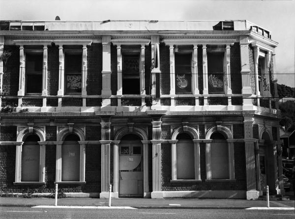 Black and white photo of Adelaide Hotel by Antony Kitchener. A brick and plaster Victorian Hotel with elaborate classical architectural details. It is unused due to earhtquake risk, the windows are boarded and covered in graffiti