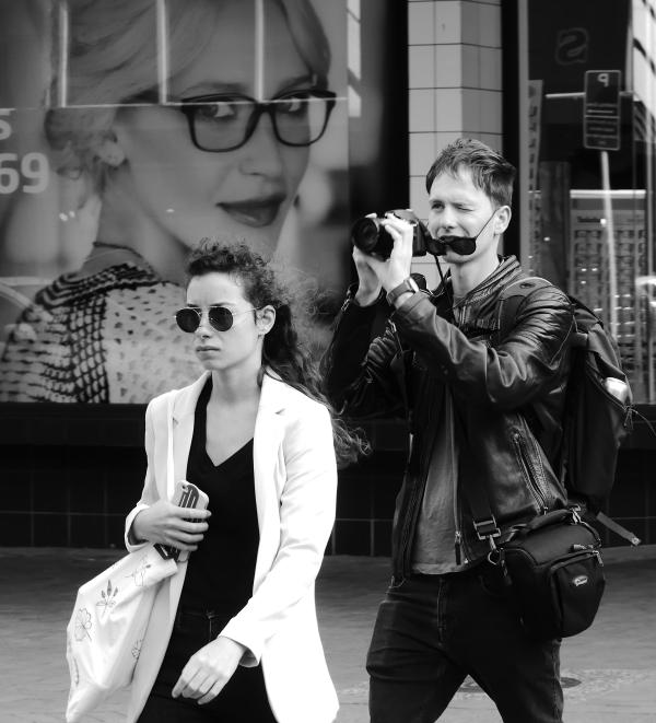Black and white photograph by Patrick Flanagan. Title 'Eyewear Abounds'. Tightly framed street scene of two pedestrians. A woman in front wears stylist sunglasses, she is followed my a male photographer raising a camera to his face to line up a shot. Behind them is a full window advertisement for an optometrist.